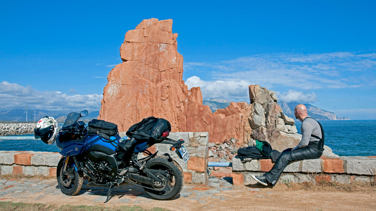 Roter Porphyrfelsen am Meer, davor ein geparktes Motorrad und ein Fahrer auf einer Mauer Roter Porphyrfelsen am Meer, davor ein geparktes Motorrad und ein Fahrer auf einer Mauer